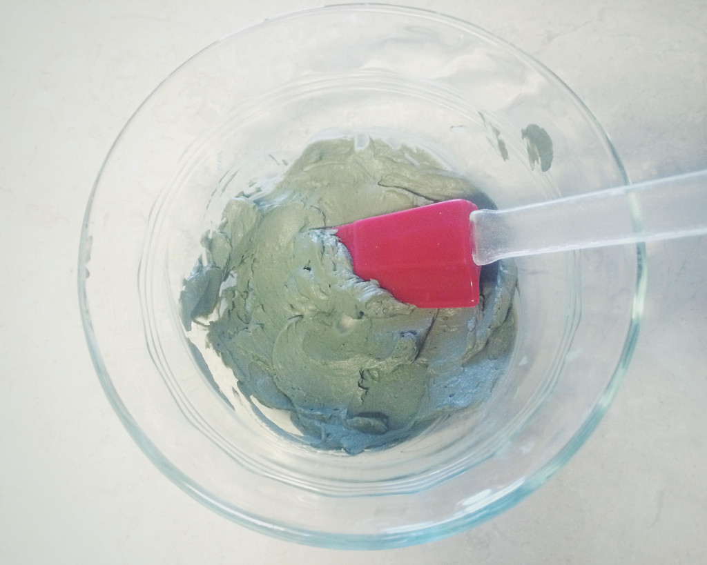 Green clay mask being mixed in glass bowl with red rubber spatula