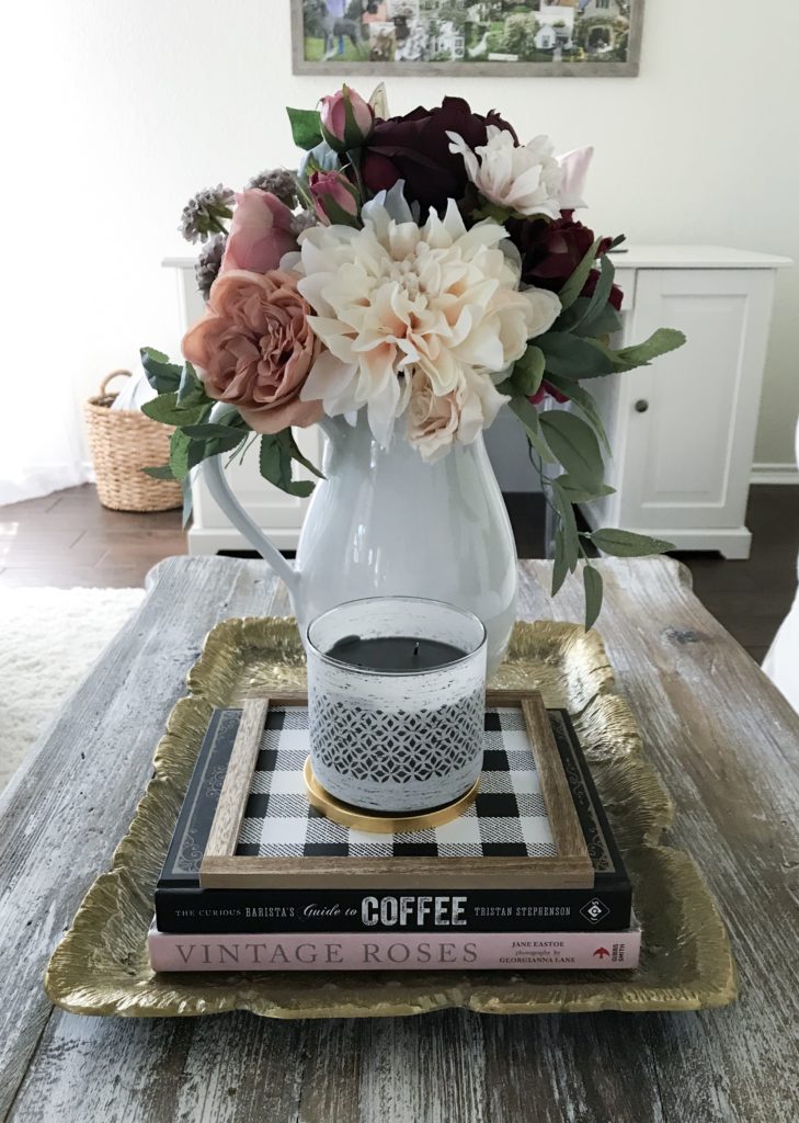 coffee table with books on gold tray and candle and white pitcher with faux flowers in it